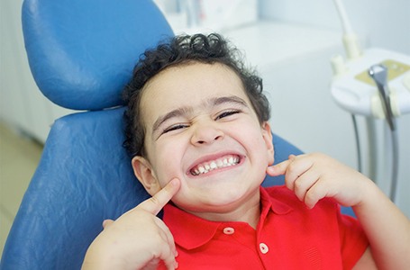 child smiling and pointing at their teeth while sitting in a dental chair